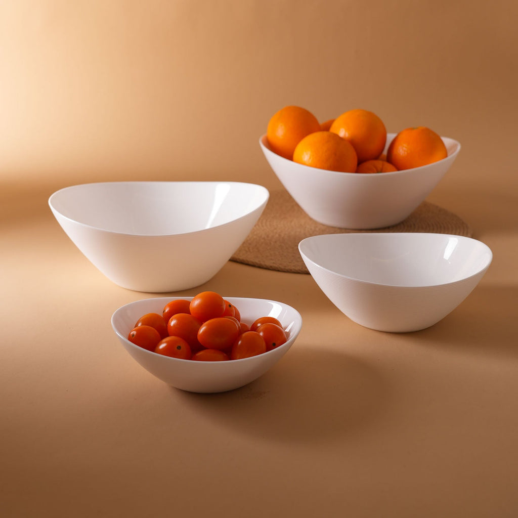 Four white opal glass bowls of varying sizes arranged on a warm beige background, with two bowls containing small orange fruits and one bowl containing red cherry tomatoes.