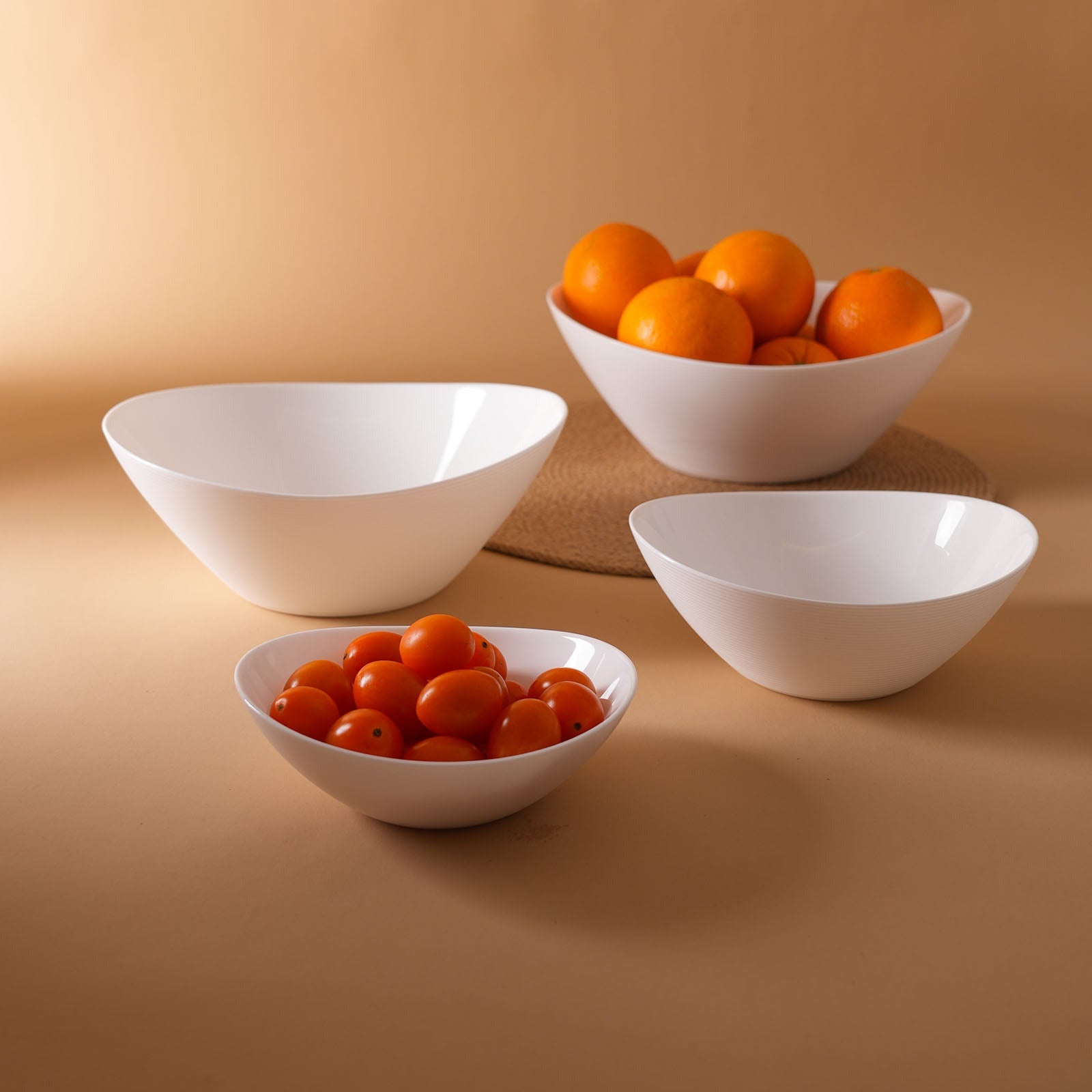 Four white opal glass bowls of varying sizes arranged on a warm beige background, with two bowls containing small orange fruits and one bowl containing red cherry tomatoes.
