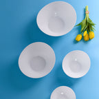 Four white opal glass bowls in different sizes arranged on a bright blue background, with a small bunch of yellow tulips in the top right.