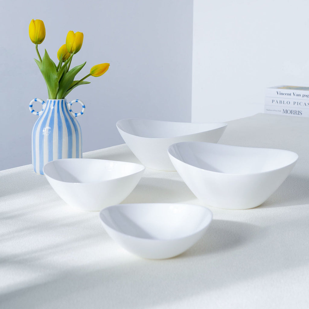 Four white oval-shaped Opal glass bowls arranged on a white surface with a blue-and-white striped vase of yellow tulips and a small book in the background.