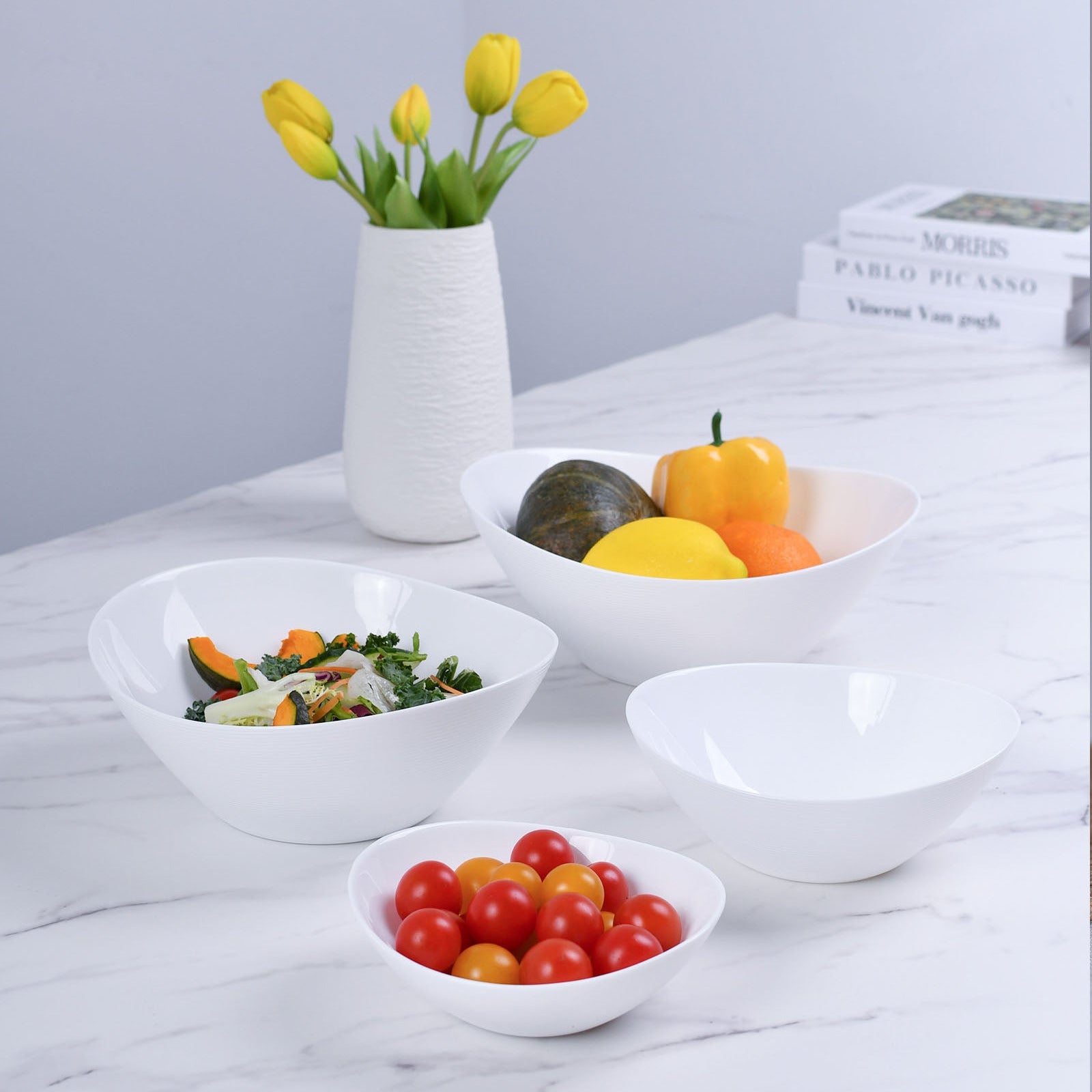 Four opal glass bowls of varying sizes arranged on a white marble surface, with one bowl holding mixed greens and vegetables, another holding yellow and orange fruits, a third bowl empty, and a fourth bowl in front holding red cherry tomatoes. A white textured vase with yellow tulips stands behind the bowls, and books are stacked in the background.