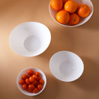 Four white opal glass bowls arranged on a warm tan background, with two bowls containing small orange fruits and tomatoes.
