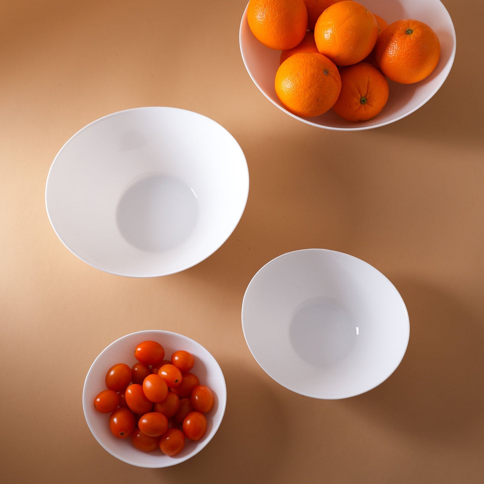 Four white opal glass bowls arranged on a warm tan background, with two bowls containing small orange fruits and tomatoes.