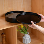 Two round black glass dinner plates on a light wood shelf, with a person's hands holding one tray and a small vase of yellow tulips below.
