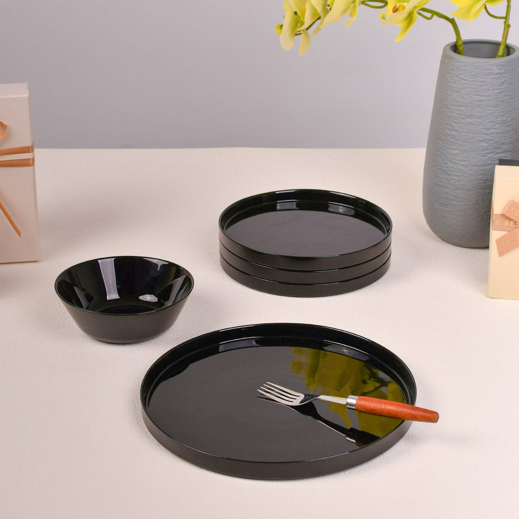 Black opal glass dinnerware set including a large platter, a matching bowl, and a stack of smaller round plates, styled on a light surface with yellow flowers in a gray vase.