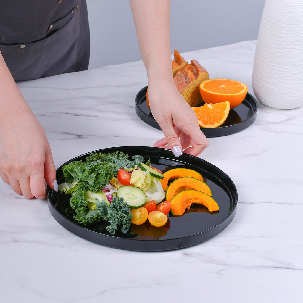 Two round black serving plates on a white marble table, with a person's hands holding the front dinner plate filled with salad and fruit slices. A second tray with food is in the background next to a white vase.