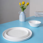 A set of white opal glass tableware, including a large plate, a smaller plate, and a bowl, arranged on a bright blue table next to a blue-and-white striped vase with yellow tulips.