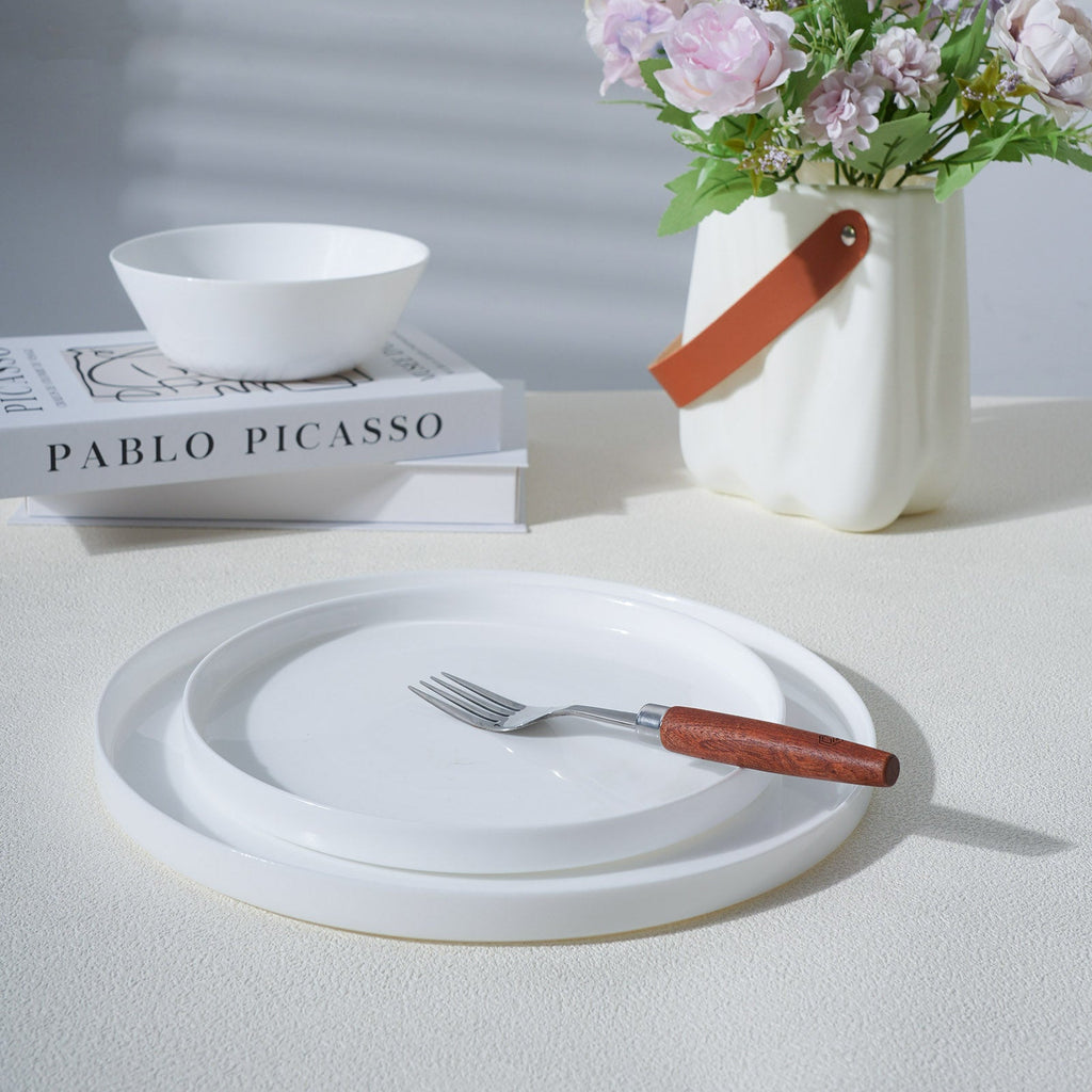 A stack of white opal glass plates with a matching white bowl, styled on a tabletop with a fork that has a brown wooden handle, next to a white vase with flowers.