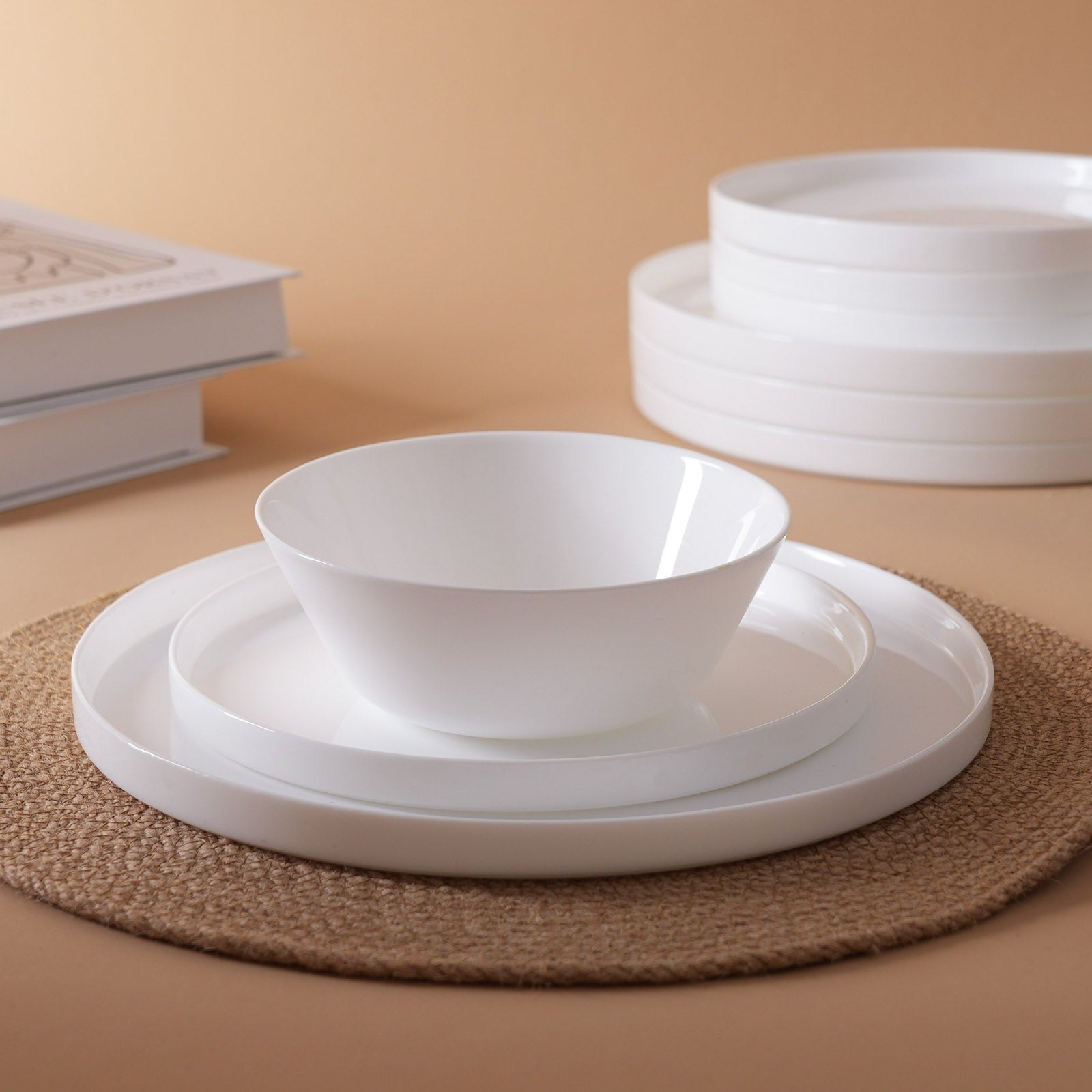 White opal glass dinnerware set including a bowl, plates, and stacked plates, arranged on a woven placemat against a warm beige background.