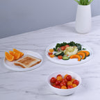Three white opal glass tableware pieces on a white marble surface: a plate with toast and orange slices, a plate with a colorful salad, and a bowl filled with cherry tomatoes. A white vase with yellow tulips sits in the background against a light gray wall.
