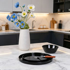 A lifestyle shot of a black opal glass serving platter with a fork and a small black bowl on a white marble kitchen island, with a white textured vase of blue and white flowers in the background.