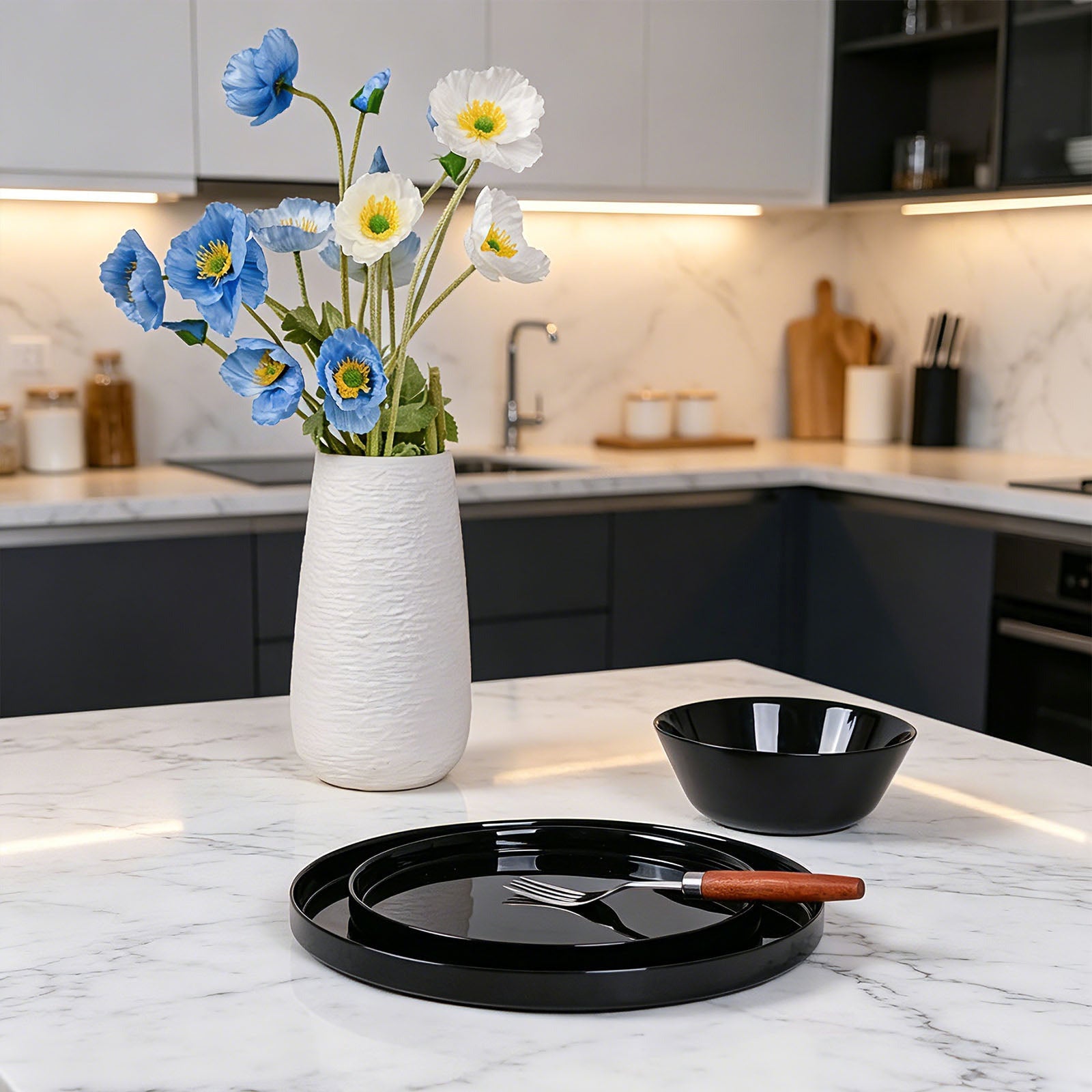 A lifestyle shot of a black opal glass serving platter with a fork and a small black bowl on a white marble kitchen island, with a white textured vase of blue and white flowers in the background.