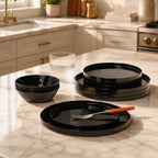 Black opal glass dinnerware set including plates and bowls arranged on a marble kitchen countertop, with a glass and a utensil in a real kitchen setting.