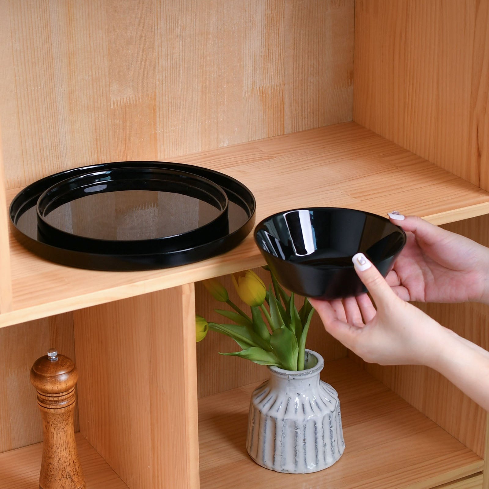 A person's hands are shown holding a small black opal glass bowl next to a larger round black tray on a light wood shelf.