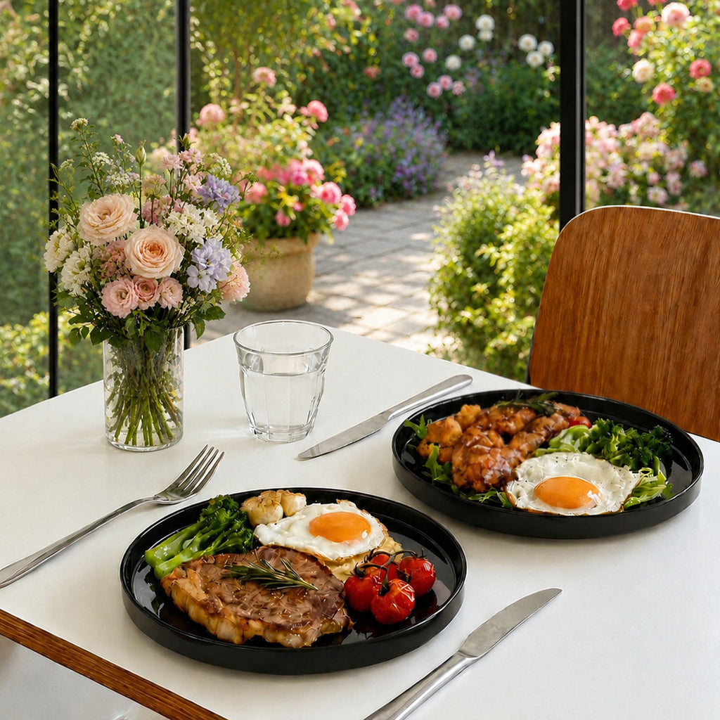 Two black opal glass plates on a white table set with cutlery, a glass of water, and a flower vase, with a bright garden patio visible through large windows in the background.