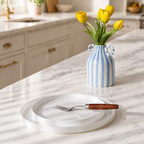 A blue and white striped ceramic vase with loop handles, filled with yellow tulips, sits on a white marble kitchen island. In the foreground, a white opal glass plate and a fork with a brown wooden handle rest on the counter.