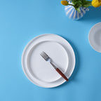 A top-down view of a white opal glass dinner plate with a fork featuring a brown wooden handle, set against a bright blue background with a striped vase of yellow tulips.