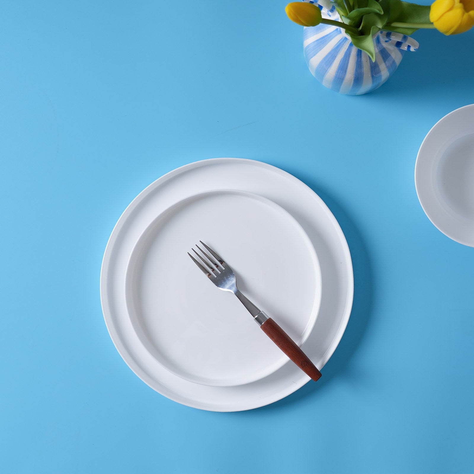 A top-down view of a white opal glass dinner plate with a fork featuring a brown wooden handle, set against a bright blue background with a striped vase of yellow tulips.