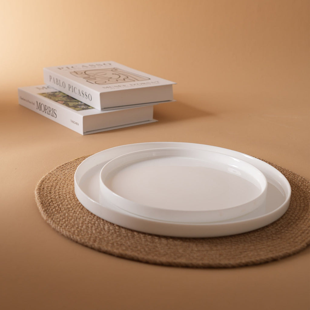 Two stacked white opal glass plates on a round woven placemat, with two books in the background against a warm beige backdrop.