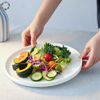 A white opal glass plate holding a colorful salad with sliced cucumbers, cherry tomatoes, kale, and pumpkin, being held by two hands over a light gray tabletop.